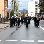Gfrupa osób idzie w pochodzie ulicami miasta. Po dwóch stronach drogi stoją budynki. Na czele pochodu idzie orkiestra.
