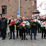Grupa mężczyzn i kobiet ubranych w odświętne stroje stoi przed budynkiem z cegłym, trzymając w rękach piękne biało-czerrowne wiązanki kwiatów.