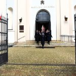 Grupa mężczyzn ubrana w odświetnie strażackie mundury wychodzi z budynku kościeła z jasną elewacją i bramą.