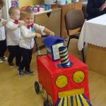 Trzech małych chłopców ubranych w białe koszule i ciemne spodnie z czerownymi muszkami prowadzi pociąg zrobiony z kartonu i wjezdzaja do sali.