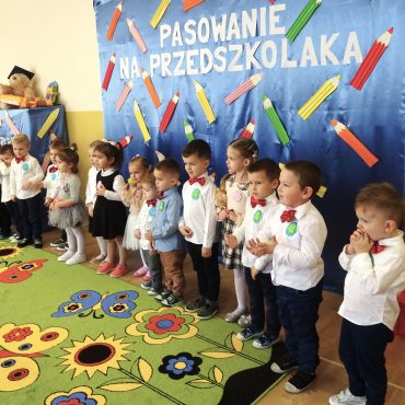 Grupa dzieci ubrana w eleganckie stroje stoi na zielonym dywanie w kolorowe kwiatki i mowią wierszyki.