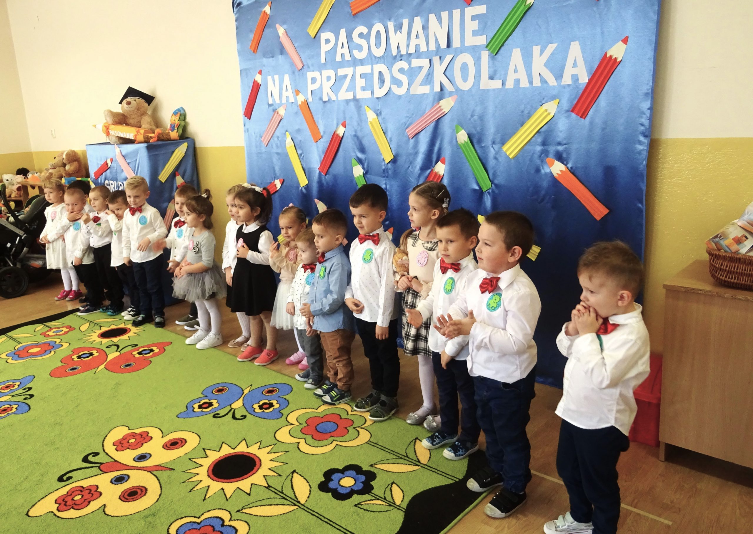 Grupa dzieci ubrana w eleganckie stroje stoi na zielonym dywanie w kolorowe kwiatki i mowią wierszyki.