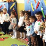 Grupa dzieci ubrana w eleganckie stroje stoi na zielonym dywanie w kolorowe kwiatki i mowią wierszyki.