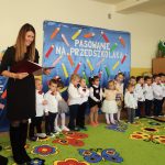 Grupa dzieci ubrana w eleganckie stroje stoi na zielonym dywanie w kolorowe kwiatki i mowią wierszyki. Przed nimi stoi kobieta o ciemnych włosach i w czarnej sukience i zapowiada występujących.