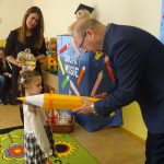 Starszy mężczyzna w gafrniturze pasuje zółtym ołowkiem dziewczynkę na przedszkolaka.