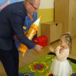 Starszy mężczyzna w gafrniturze pasuje zółtym ołowkiem dziewczynkę na przedszkolaka