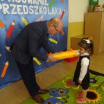 Starszy mężczyzna w gafrniturze pasuje zółtym ołowkiem dziewczynkę na przedszkolaka