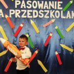 Chłopiec ubrany w białą koszulę stoi w sali i trzyma duży ołówek. Za nim stoi ścianka koloru granatowego ozdobiona kolorowymi ołowkami wycitymi z piaeru i napisem Pasowanie na Przedszkolaka./