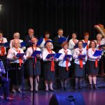 Grupa starszych osób kobiet i mężczyzn ubranych w odświętne stroje białe koszule spiewają na scenie kina. W rękach trzymają granatowe śpiewniki. Na szyi zawieszone mają czerwone wstążki.