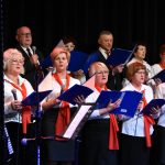 Grupa starszych osób kobiet i mężczyzn ubranych w odświętne stroje białe koszule spiewają na scenie kina. W rękach trzymają granatowe śpiewniki. Na szyi zawieszone mają czerwone wstążki.