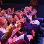 Grupa osób siedzi na fotelach w kinie i bije brawo, ogladając występ dzieci.