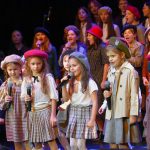 Grupa dzieci w odświętnych strojach stoi na scenie i spiewa pieśni patriotyczne.