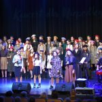 Grupa dzieci w odświętnych strojach stoi na scenie i spiewa pieśni patriotyczne.