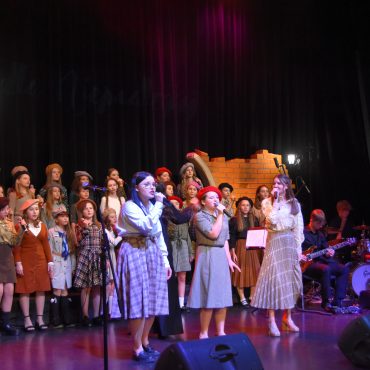 Grupa dzieci w odświętnych strojach stoi na scenie i spiewa pieśni patriotyczne. Na pierwszym planie śpiewają trzy dziewczyny.