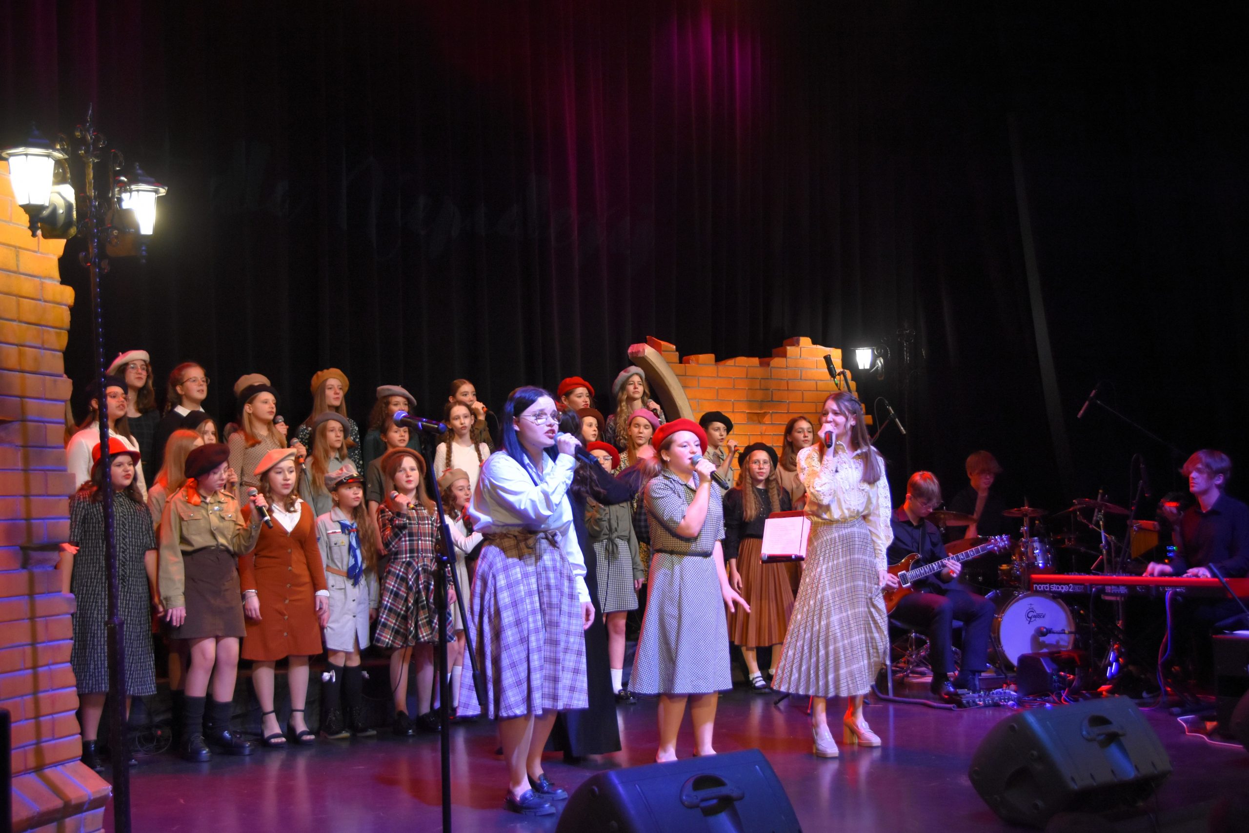 Grupa dzieci w odświętnych strojach stoi na scenie i spiewa pieśni patriotyczne. Na pierwszym planie śpiewają trzy dziewczyny.