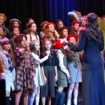 Grupa dzieci w odświętnych strojach stoi na scenie i spiewa pieśni patriotyczne.