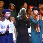 Grupa dzieci w odświętnych strojach stoi na scenie i spiewa pieśni patriotyczne. Na pierwszym planie stoją dwie młode dziewczyny ubrane w białą koszulę i zieloną sukienkę i śpiewają do mikrofonu.