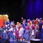 Grupa dzieci w odświętnych strojach stoi na scenie i spiewa pieśni patriotyczne. Dzieci trzymają w rękach falgi małe biało-czerwone.