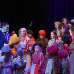 Grupa dzieci w odświętnych strojach stoi na scenie i spiewa pieśni patriotyczne. Obok na scenie stoi mężczyzna w garniturze i przemawia do mikrofonu. Obok stoi Młoda kobieta o blond włosach i się usmiecha.