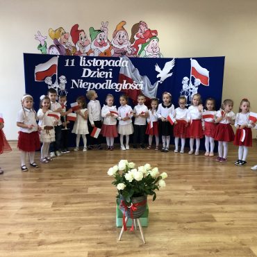 Grupa dzieci ubranych w białe bluzki i element czerownego stoją na środku sali i mówią iwerszyki. Za nimi stoi tablica przesłoniona granatowym płótnem, ozdobiona biało-czerwonymi elementami polskości. Na środku stoi wazon z białymi różami.