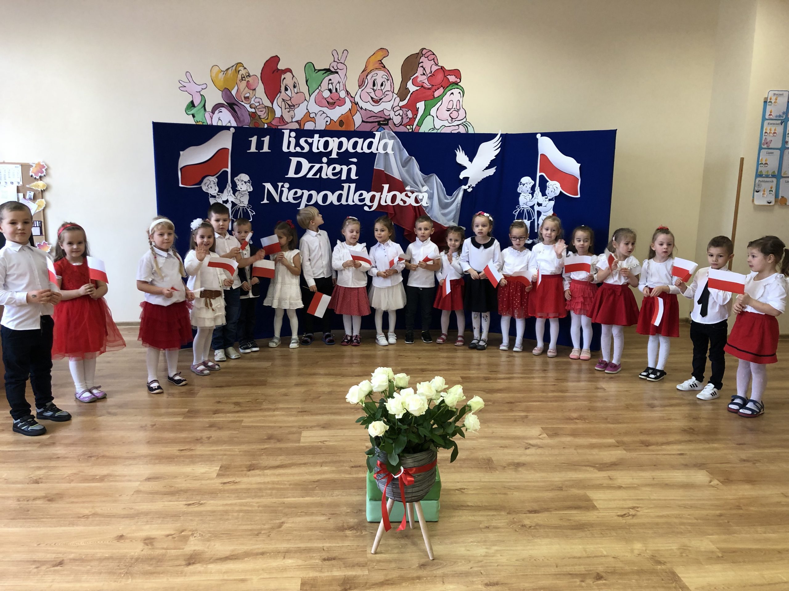 Grupa dzieci ubranych w białe bluzki i element czerownego stoją na środku sali i mówią iwerszyki. Za nimi stoi tablica przesłoniona granatowym płótnem, ozdobiona biało-czerwonymi elementami polskości. Na środku stoi wazon z białymi różami.