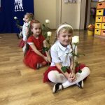 Trzy dziewczynki siedzą na parkiecie w sali przedszkolnej, ubrane w odświętne biało czerwone stroje, w rękach trzymają białą różyczkę.