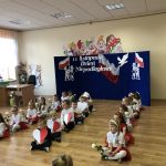 Grupa dzieci ubranych w białe i czeerwone stroje tańczą na sali przedszkolnej z kwiatami serduszkami wyciętymi z papieru.