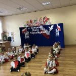 Grupa dzieci ubranych w białe i czeerwone stroje tańczą na sali przedszkolnej z kwiatami serduszkami wyciętymi z papieru.