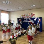 Grupa dzieci ubranych w białe i czeerwone stroje tańczą na sali przedszkolnej z kwiatami.