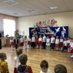 Grupa dzieci ubranych w białe bluzki i element czerownego stoją na środku sali i mówią iwerszyki. Za nimi stoi tablica przesłoniona granatowym płótnem, ozdobiona biało-czerwonymi elementami polskości.