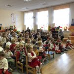 Grupa dzieci odświetnie ubranych siedzi na krzesłach w dużej sali przedszkolnej i ogląda przedstawienie.