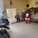 Występ starszej kobiety grającej na gitarze oraz męczyzny przebranego w stroju swietego mikołaja, osoby na wozkach inwaldzkich obserwują występ.