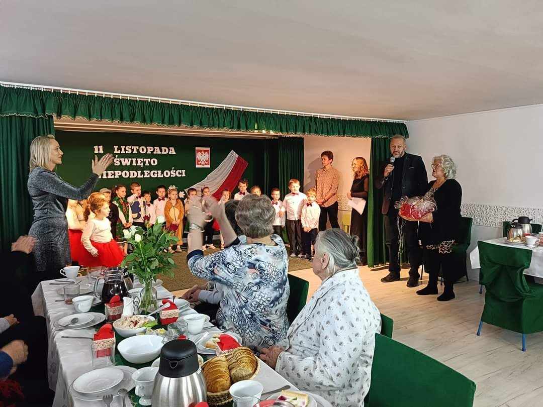 Grupa starszych kobiet siedzi przy stole swiatecznym i smieją się do siebie oraz ogladają występ dzieci.