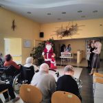 Występ starszej kobiety grającej na gitarze oraz męczyzny przebranego w stroju swietego mikołaja, osoby na wozkach inwaldzkich obserwują występ.