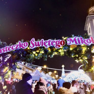 napis miasteczko swietego mikołaja na stelazu wyłożonym girlanda na tle buydnku ratusza z jasna elewacją.