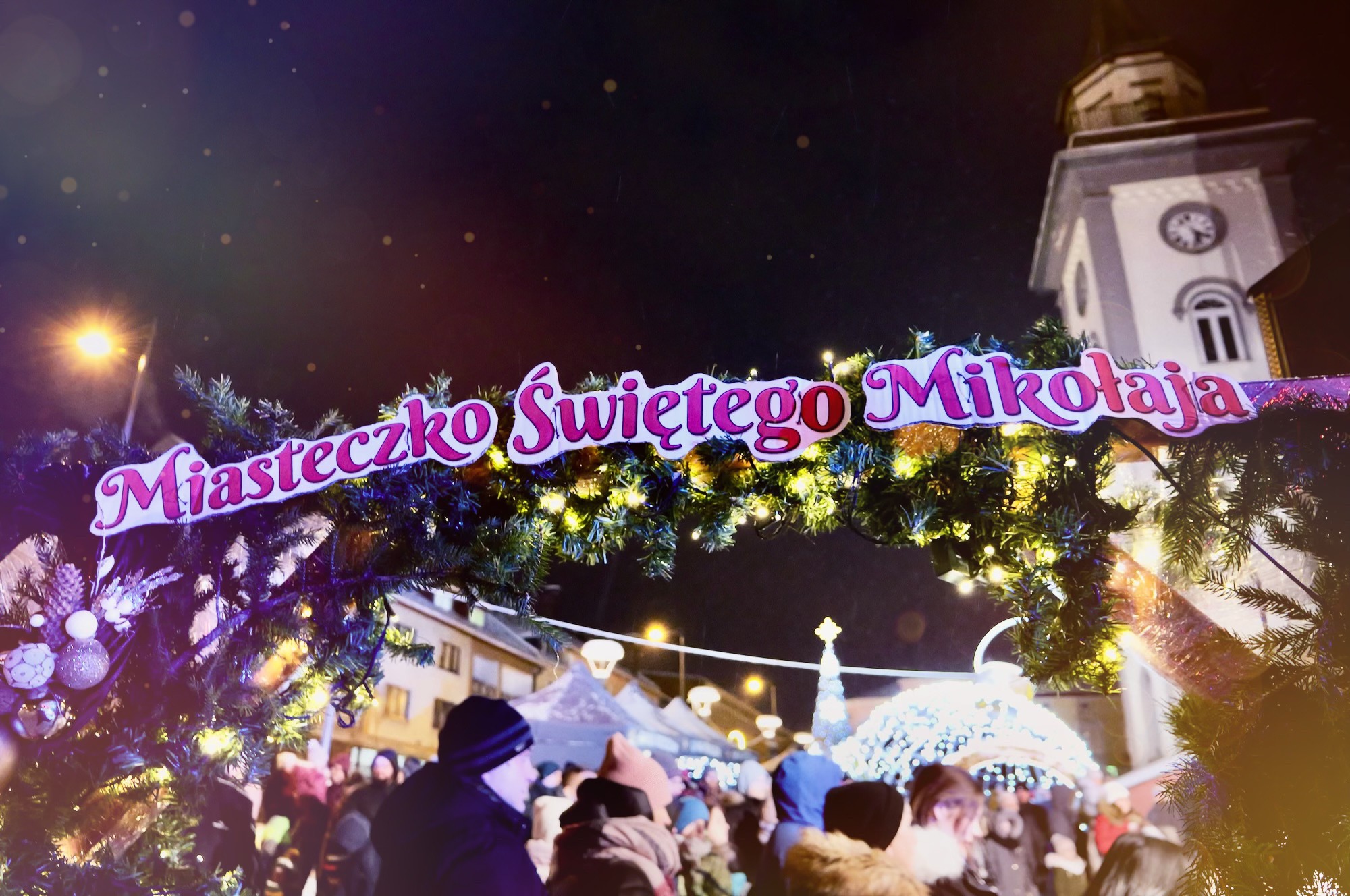 napis miasteczko swietego mikołaja na stelazu wyłożonym girlanda na tle buydnku ratusza z jasna elewacją.