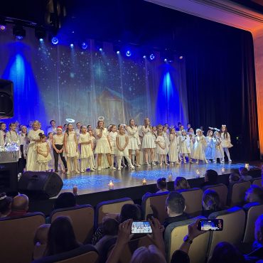 Grupa dzieci i młodzieży ubranych w białe stroje stoją na scenie tanecznej i spiewają koledy. Przed scena na fotelach kinowych siedzą ludzie i patrzą na wystep.