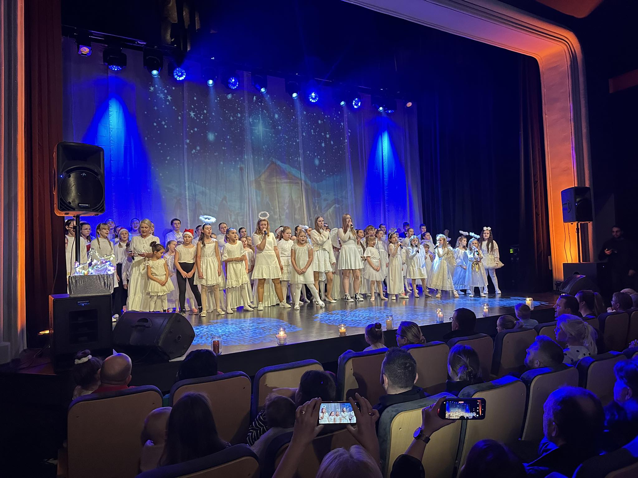 Grupa dzieci i młodzieży ubranych w białe stroje stoją na scenie tanecznej i spiewają koledy. Przed scena na fotelach kinowych siedzą ludzie i patrzą na wystep.