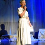 Kobieta w srednim wieku o krótkich blond włosach ubrana w białą sukienkę długę stoi na scenie kina i przemawia do mikrofonu.