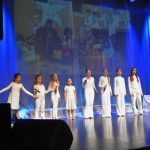 Grupa dziewczynek ubranych na biało śpiewa na scenie kina.