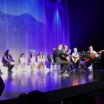 Grupa dzieci i młodzieży siedzi na scenie kina i gra na instrumentach.