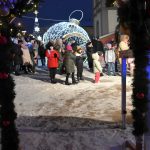 Grupa ludzi stoi na płycie rynku miasta. Na rynku stoi owietlona swiatelkami duza bombka i choinka.