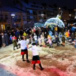 na płycie ryunku grupa dzieci bawi się z kobietami przebranymi za elfów. Zabawe obserwują osoby starsze i rodzice.