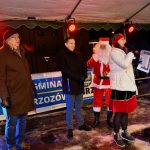 Na scenie stoi siwety mikołaj w czerownym stroju oraz dwóch mężczyzn ubranych w zimowe kurtki oraz kobieta w stroju elfa przemawia do mikrofonu.