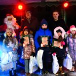 Piątka dzieci ubranych w zimowe stroje stoi na scenie, za nimi dwoch meżczzyn oraz swiety mikołaj pozują do zdjecia.