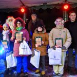 Piątka dzieci ubranych w zimowe stroje stoi na scenie, za nimi dwoch meżczzyn oraz swiety mikołaj pozują do zdjecia.