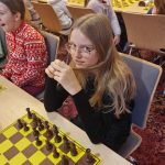 Dziewczynka w długich włosach siedzi przy tablicy szachowej i gra w szachy. Obok niej siedzą inne dzieci.