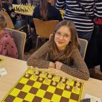 Dziewczynka w długich włosach i okularach siedzi przy tablicy szachowej i gra w szachy. Obok niej siedzą inne dzieci.
