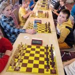 Grupa dzieci siedzi za stołem na którym są ułożone szachy.