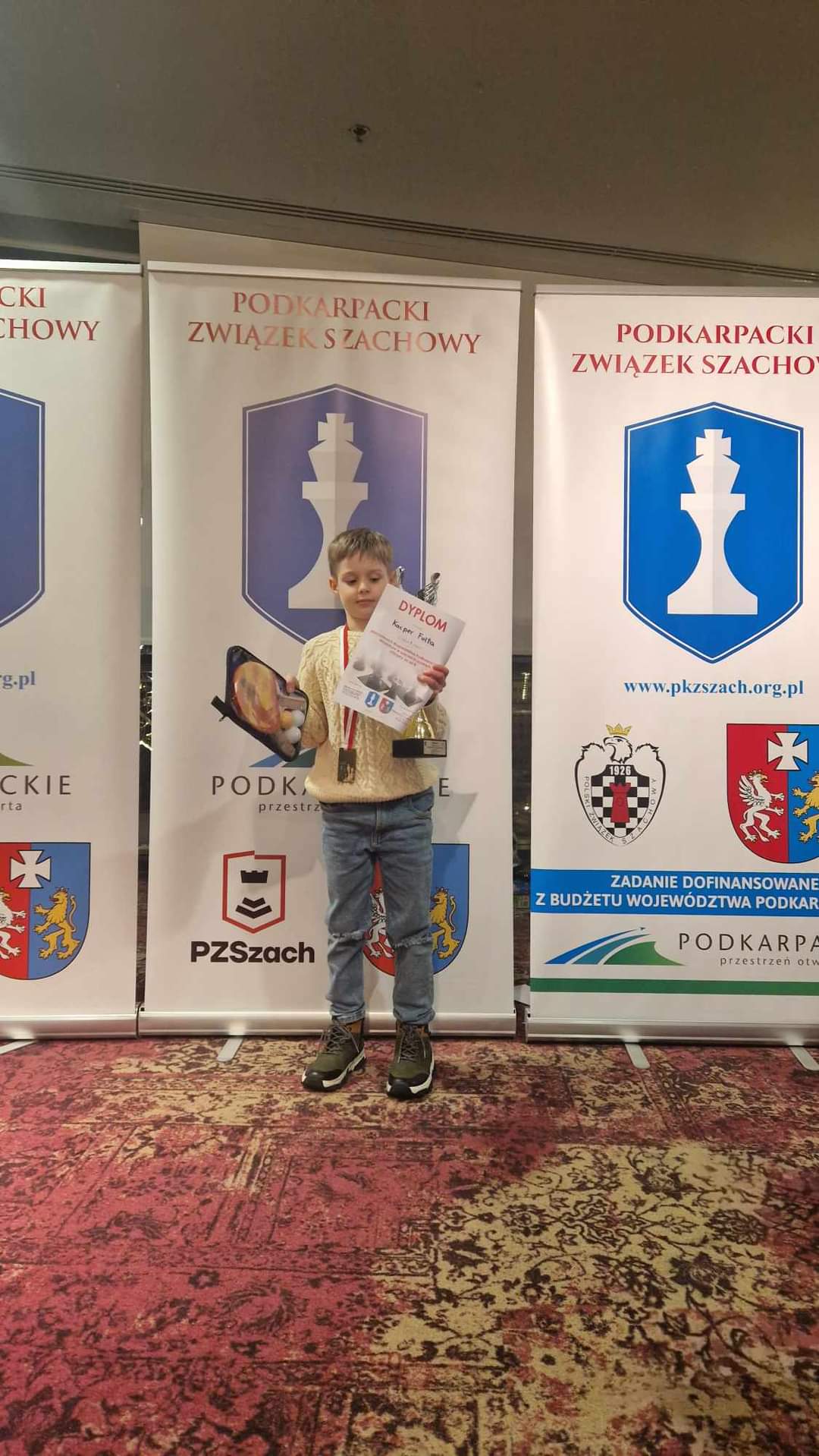 Chłopiec stoi na podium z dyplomem, medalem i pucharem.